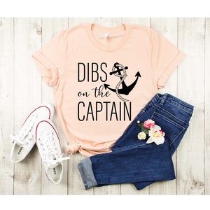 May include: A peach-colored t-shirt with the text "DIBS on the CAPTAIN" and a black anchor with a rope design. The shirt is laid out on a wooden surface with a pair of white sneakers and blue jeans.