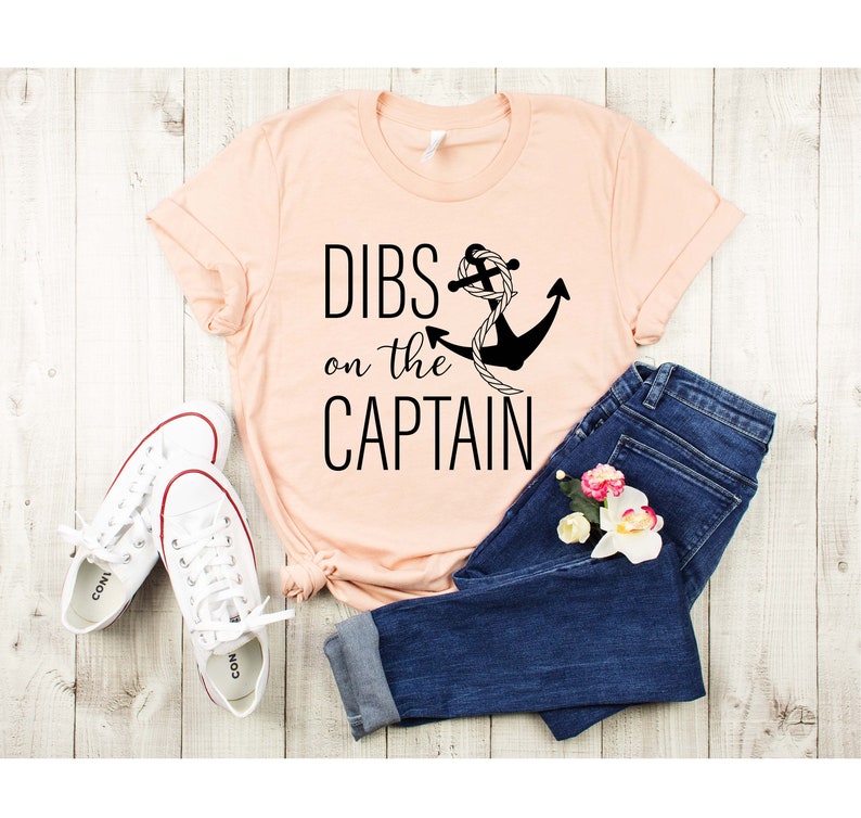 May include: A peach-colored t-shirt with the text "DIBS on the CAPTAIN" and a black anchor with a rope design. The shirt is laid out on a wooden surface with a pair of white sneakers and blue jeans.