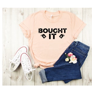 Puede incluir: Una camiseta de color melocot&oacute;n con el texto "BOUGHT IT" en letras negras y dos etiquetas de precio negras con signos de d&oacute;lar. La camiseta est&aacute; colocada sobre una superficie de madera clara con un par de zapatillas blancas y unos vaqueros azules.