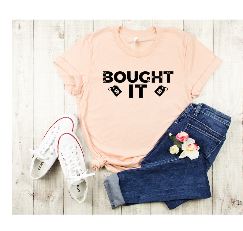Puede incluir: Una camiseta de color melocot&oacute;n con el texto "BOUGHT IT" en letras negras y dos etiquetas de precio negras con signos de d&oacute;lar. La camiseta est&aacute; colocada sobre una superficie de madera clara con un par de zapatillas blancas y unos vaqueros azules.