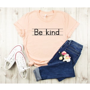 Puede incluir: Una camiseta de color melocot&oacute;n con el texto "Be kind" escrito en negro sobre fondo blanco. La camiseta se combina con unos vaqueros azules y zapatillas blancas.