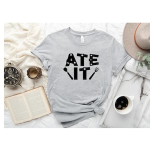 Puede incluir: Una camiseta gris con el texto "ATE IT" en letras negras. Las letras est&aacute;n formadas por im&aacute;genes de utensilios de cocina, como una cuchara, un cuchillo y un tenedor.