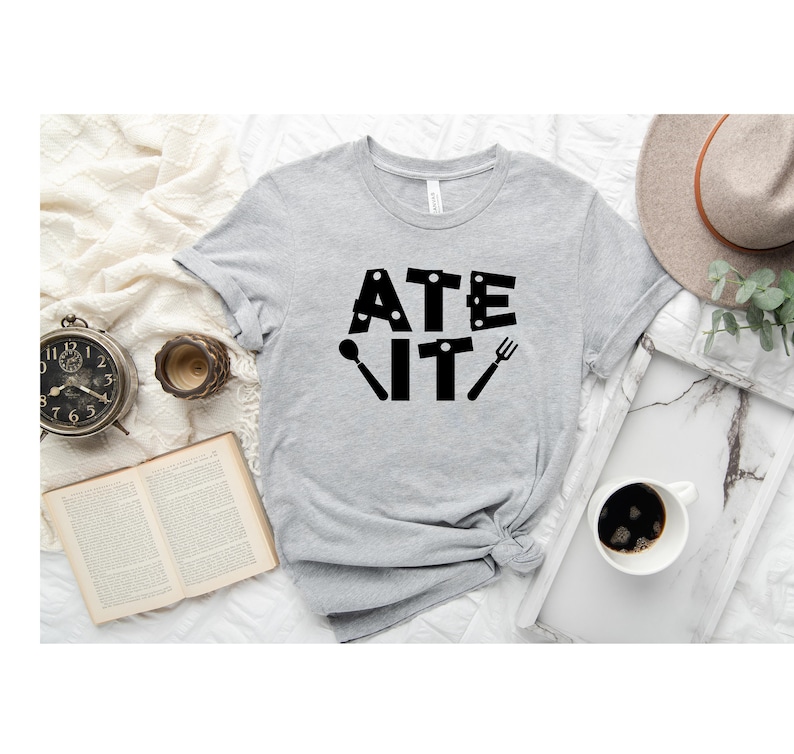 Puede incluir: Una camiseta gris con el texto "ATE IT" en letras negras. Las letras est&aacute;n formadas por im&aacute;genes de utensilios de cocina, como una cuchara, un cuchillo y un tenedor.
