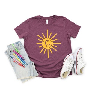 Pode incluir: Uma camiseta de manga curta cor vinho com um gr&aacute;fico de sol amarelo. O sol &eacute; desenhado em um estilo simples e infantil. A camiseta est&aacute; colocada sobre um fundo branco com um par de t&ecirc;nis brancos e um par de shorts jeans com l&aacute;pis de cor.