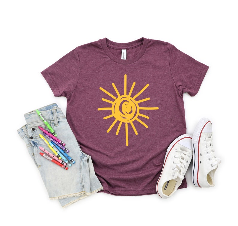 Pode incluir: Uma camiseta de manga curta cor vinho com um gr&aacute;fico de sol amarelo. O sol &eacute; desenhado em um estilo simples e infantil. A camiseta est&aacute; colocada sobre um fundo branco com um par de t&ecirc;nis brancos e um par de shorts jeans com l&aacute;pis de cor.