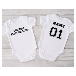 May include: Two white short-sleeved baby bodysuits. The left bodysuit features the black text CUSTOM TEXT OR LOGO. The right bodysuit displays the black text NAME above the large black number 01 with a white outline and black border.