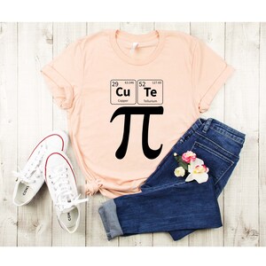 May include: A peach-colored t-shirt with a black graphic of the mathematical symbol pi and the chemical symbols for copper and tellurium. The shirt is laid out on a wooden surface with a pair of white sneakers and blue jeans.
