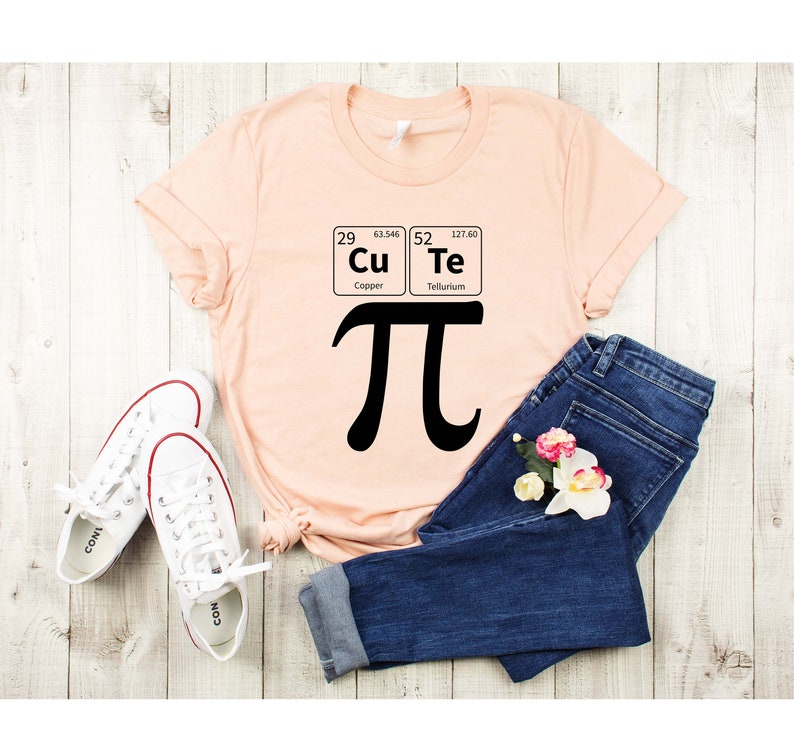 May include: A peach-colored t-shirt with a black graphic of the mathematical symbol pi and the chemical symbols for copper and tellurium. The shirt is laid out on a wooden surface with a pair of white sneakers and blue jeans.
