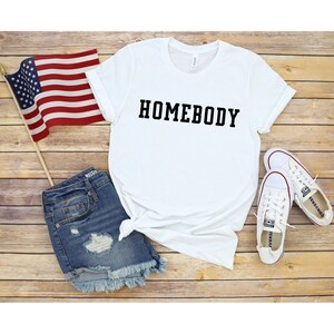 May include: White t-shirt with the text "HOMEBODY" printed in black on the front. The shirt is laid out on a wooden surface with a pair of white sneakers, denim shorts, and an American flag.