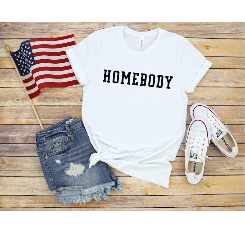May include: White t-shirt with the text "HOMEBODY" printed in black on the front. The shirt is laid out on a wooden surface with a pair of white sneakers, denim shorts, and an American flag.