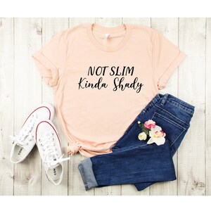 Puede incluir: Una camiseta de color melocot&oacute;n con el texto "Not Slim Kinda Shady" en letras negras. La camiseta est&aacute; colocada sobre una superficie de madera blanca con un par de zapatillas blancas y unos vaqueros azules.