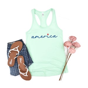 May include: A light green tank top with the word "america" in blue script with a red star above the "a". The tank top is lying flat on a white background with a pair of brown flip flops and a pair of blue denim shorts with a white floral pattern.