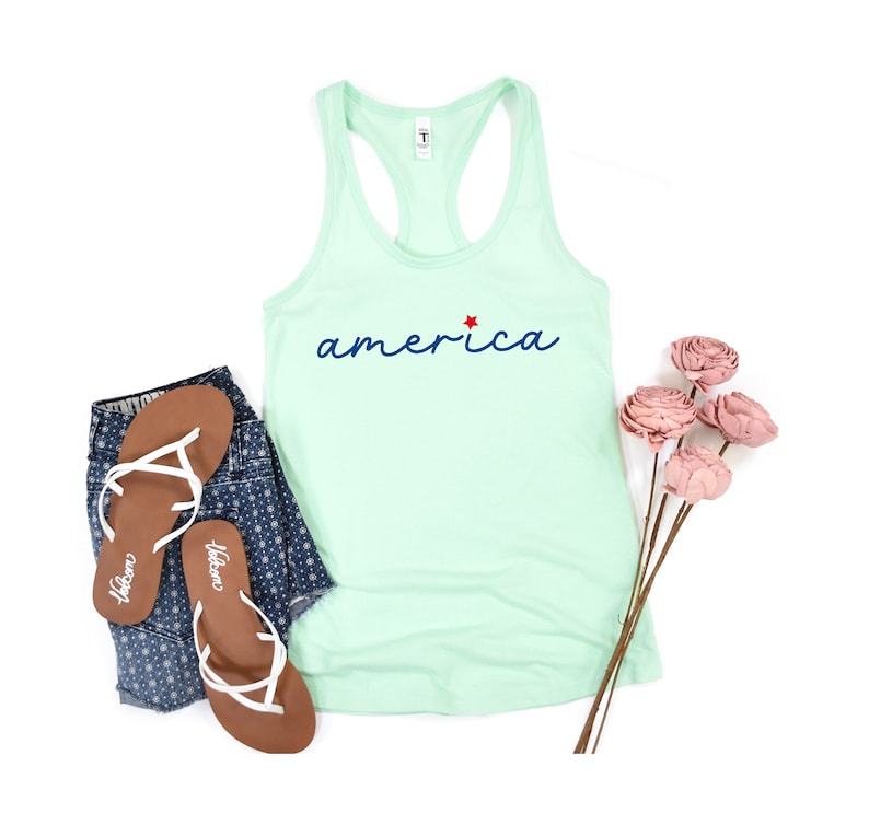 May include: A light green tank top with the word "america" in blue script with a red star above the "a". The tank top is lying flat on a white background with a pair of brown flip flops and a pair of blue denim shorts with a white floral pattern.