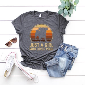 Peut inclure: Un t-shirt gris fonc&eacute; avec un graphique r&eacute;tro d'un carlin devant un coucher de soleil. Le texte "Just a girl who loves pugs" est imprim&eacute; sous le graphique.