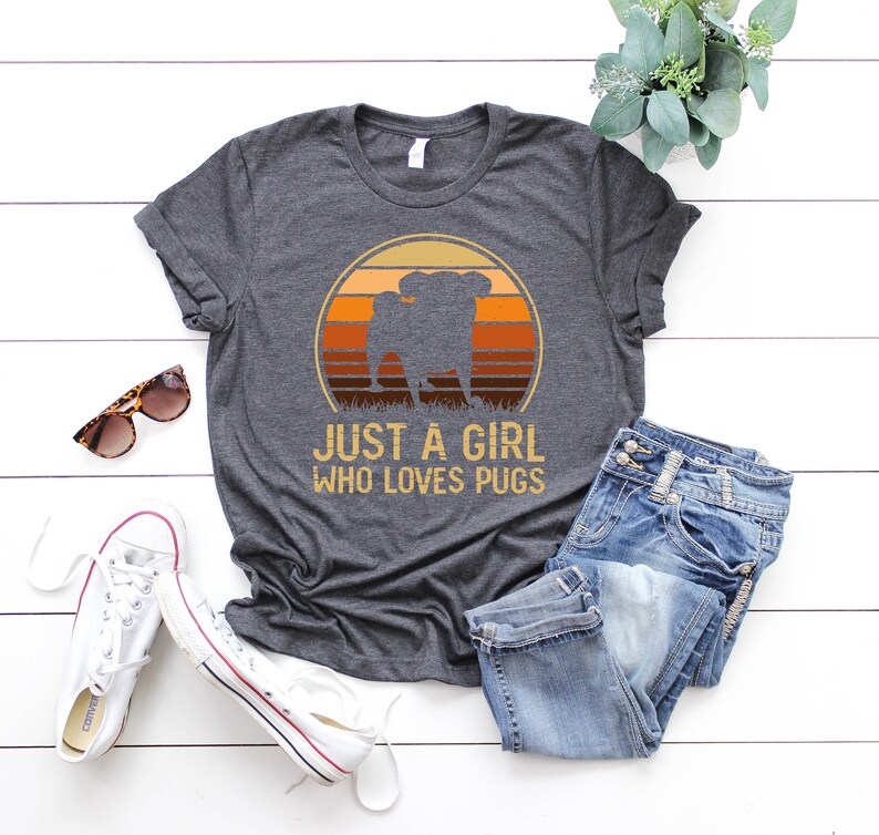 Peut inclure: Un t-shirt gris fonc&eacute; avec un graphique r&eacute;tro d'un carlin devant un coucher de soleil. Le texte "Just a girl who loves pugs" est imprim&eacute; sous le graphique.