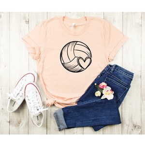 May include: A peach-colored t-shirt with a black and white illustration of a volleyball with a heart on it. The shirt is laid out on a light wood surface with a pair of white sneakers and blue jeans.