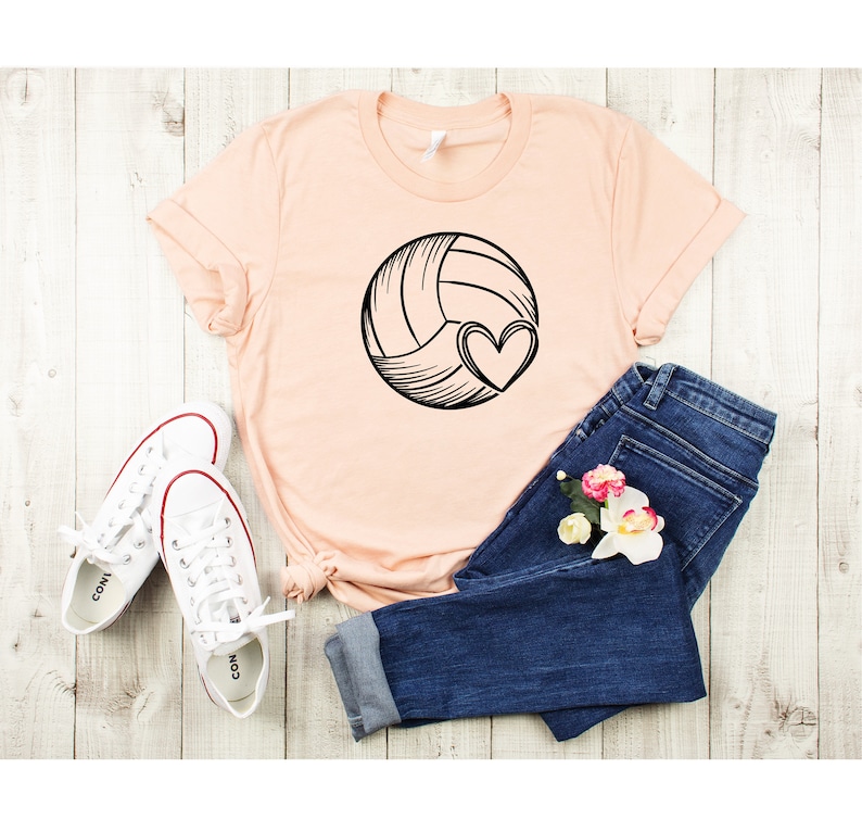 May include: A peach-colored t-shirt with a black and white illustration of a volleyball with a heart on it. The shirt is laid out on a light wood surface with a pair of white sneakers and blue jeans.