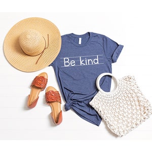 Puede incluir: Una camiseta azul con el texto "Be Kind" escrito en blanco. La camiseta est&aacute; colocada sobre una superficie de madera blanca con un sombrero de paja, dos sandalias marrones y un bolso de crochet blanco.