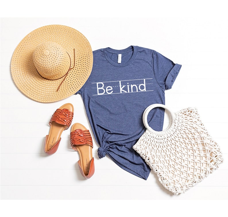 Puede incluir: Una camiseta azul con el texto "Be Kind" escrito en blanco. La camiseta est&aacute; colocada sobre una superficie de madera blanca con un sombrero de paja, dos sandalias marrones y un bolso de crochet blanco.