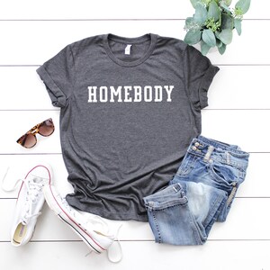 May include: A dark gray t-shirt with the word "HOMEBODY" printed in white on the front. The shirt is laid flat on a white wooden surface with a pair of white sneakers, a pair of sunglasses, and a pair of blue jeans.
