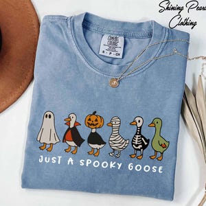 A blue t-shirt with a Halloween-themed graphic of a group of cartoon ducks dressed in costumes, accompanied by the text "Just a spooky goose".
