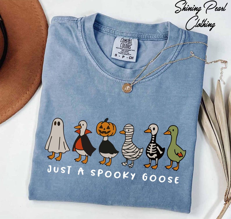 A blue t-shirt with a Halloween-themed graphic of a group of cartoon ducks dressed in costumes, accompanied by the text "Just a spooky goose".