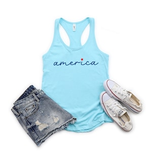 May include: A light blue tank top with the word "america" in blue script with a red star above the "a". The tank top is paired with denim shorts and white trainers.