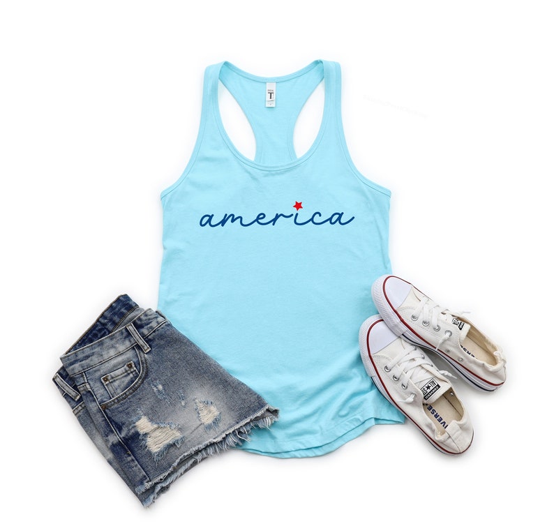May include: A light blue tank top with the word "america" in blue script with a red star above the "a". The tank top is paired with denim shorts and white trainers.