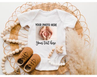 May include: A white short-sleeved baby bodysuit featuring a rectangular graphic with the text YOUR PHOTO HERE above an image of hands holding baby feet, and the text Your text here printed in black script below the graphic.