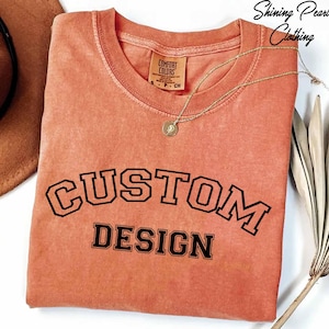 a pink t-shirt with the text "CUSTOM DESIGN" printed on it, along with a necklace and a hat.