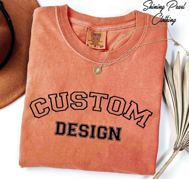 a pink t-shirt with the text "CUSTOM DESIGN" printed on it, along with a necklace and a hat.