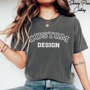 A person wearing a grey t-shirt with the text "CUSTOM DESIGN" printed on it, along with a necklace and bracelets.