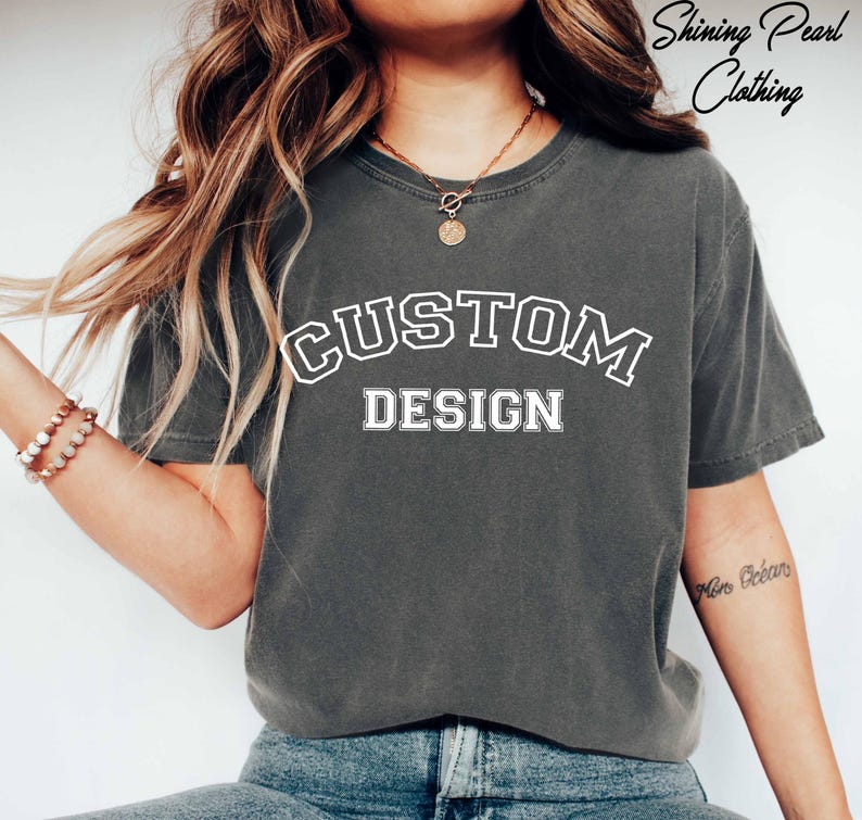 A person wearing a grey t-shirt with the text "CUSTOM DESIGN" printed on it, along with a necklace and bracelets.