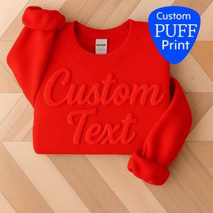 May include: A bright red sweatshirt with the words "Custom Text" in a raised, textured font. The sleeves are cuffed. A blue, shield-shaped graphic in the upper right corner reads "Custom PUFF Print".