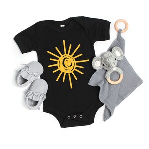 Pode incluir: Um macac&atilde;o preto para beb&ecirc; com um gr&aacute;fico de sol amarelo. O macac&atilde;o est&aacute; sobre um fundo branco com um chocalho de elefante cinza e um anel de madeira.