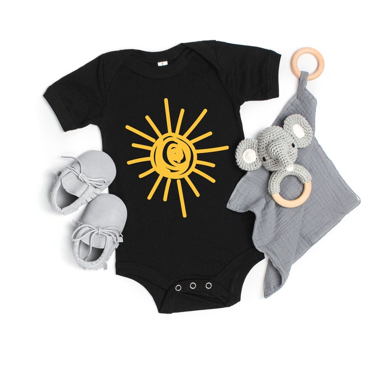Pode incluir: Um macac&atilde;o preto para beb&ecirc; com um gr&aacute;fico de sol amarelo. O macac&atilde;o est&aacute; sobre um fundo branco com um chocalho de elefante cinza e um anel de madeira.