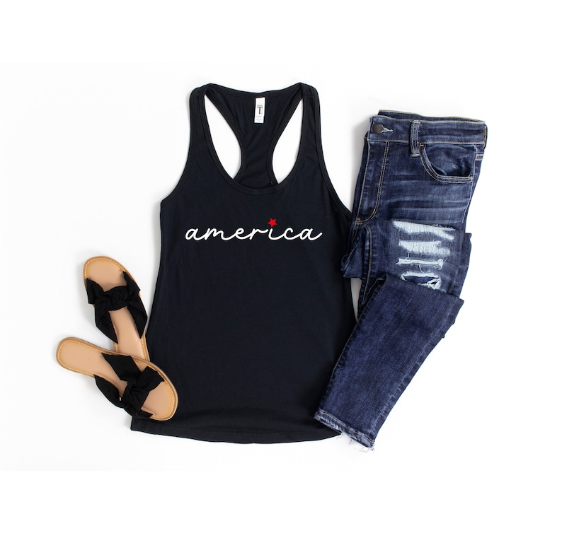 May include: A black tank top with a white cursive inscription that reads "america" with a small red star above the "a". The tank top is paired with blue jeans and brown sandals.