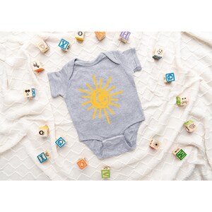 Pode incluir: Um macac&atilde;o de beb&ecirc; cinza com um gr&aacute;fico de sol amarelo. O macac&atilde;o &eacute; cercado por blocos de madeira com letras.