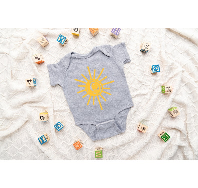Pode incluir: Um macac&atilde;o de beb&ecirc; cinza com um gr&aacute;fico de sol amarelo. O macac&atilde;o &eacute; cercado por blocos de madeira com letras.