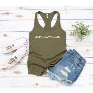 May include: A green tank top with the word "america" written in white cursive with a red star above the "a". The tank top is lying on a white wooden surface with a pair of white trainers and blue denim shorts.
