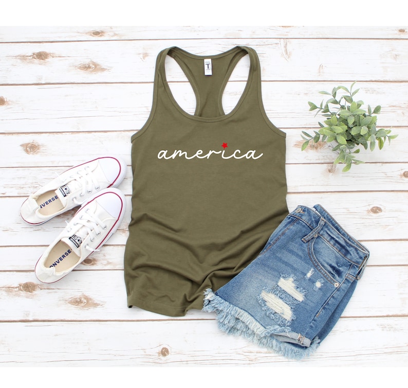 May include: A green tank top with the word "america" written in white cursive with a red star above the "a". The tank top is lying on a white wooden surface with a pair of white trainers and blue denim shorts.