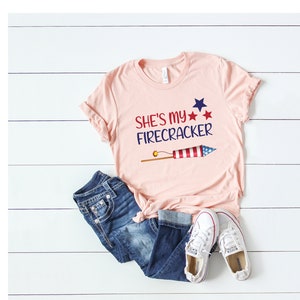 May include: A pink t-shirt with the text "She's my Firecracker" and a red, white and blue firework graphic. The shirt is paired with blue jeans and white trainers.
