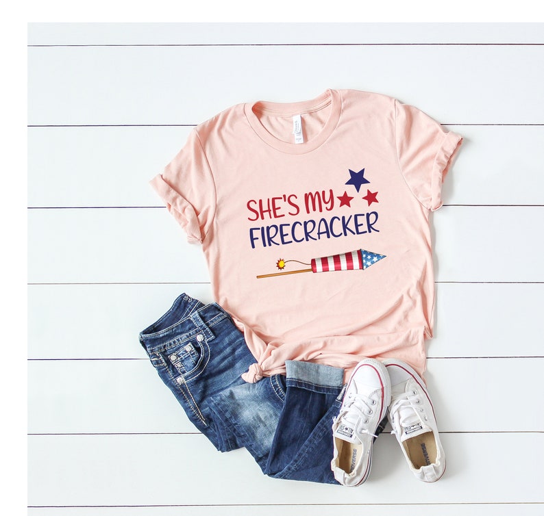 May include: A pink t-shirt with the text "She's my Firecracker" and a red, white and blue firework graphic. The shirt is paired with blue jeans and white trainers.