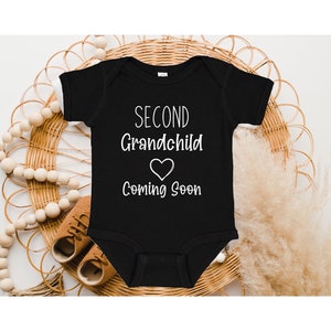 May include: Black baby onesie with white text that reads "Second Grandchild Coming Soon" with a heart symbol.