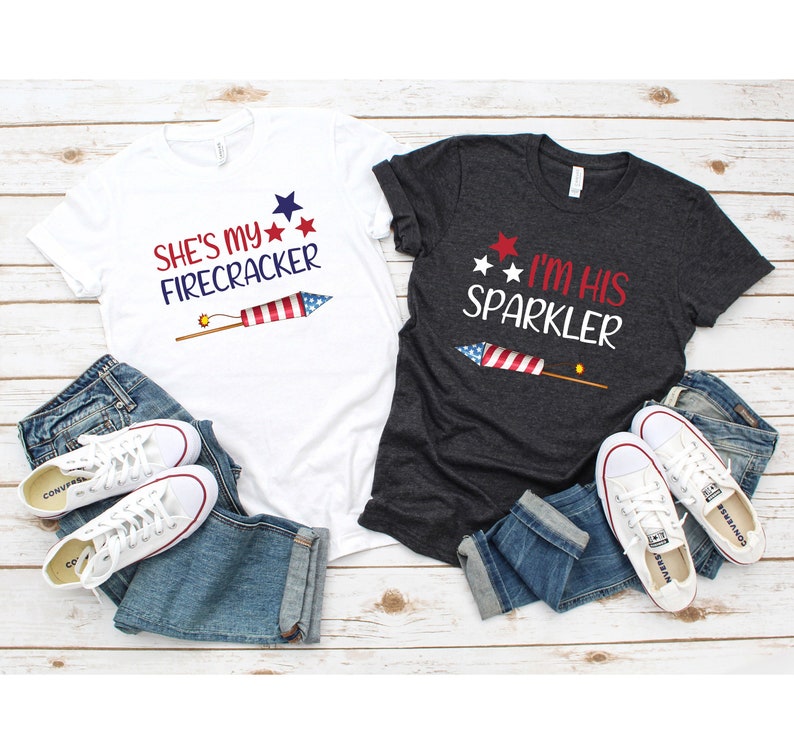 May include: Two t-shirts with a Fourth of July theme. The white t-shirt says "She's my Firecracker" with a red, white and blue firework graphic. The dark grey t-shirt says "I'm his Sparkler" with a red, white and blue sparkler graphic.