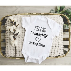 May include: White baby onesie with black text that reads "Second Grandchild Coming Soon" with a heart symbol.