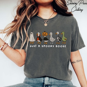 A woman wearing a gray t-shirt with a graphic design of a group of cartoon-style geese, including a ghost, skeleton, and mummy goose, along with the text "Just a Spooky Goose".