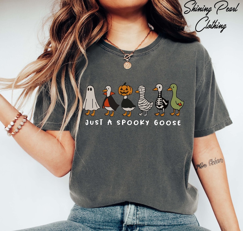 A woman wearing a gray t-shirt with a graphic design of a group of cartoon-style geese, including a ghost, skeleton, and mummy goose, along with the text "Just a Spooky Goose".