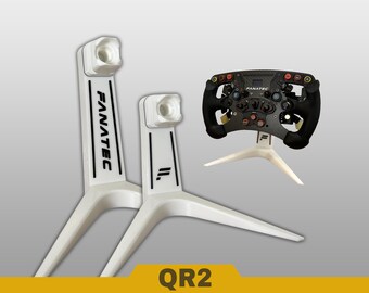 May include: Two white stands for racing wheels, each featuring a vertical column with a top mounting point and a wide tripod base. One stand displays the text FANATEC, the other a logo. A black racing steering wheel is mounted on a third stand.