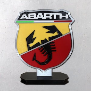 May include: A shield-shaped Abarth emblem sign featuring a black scorpion graphic centered over a yellow and red background. The top section displays the text ABARTH in white letters on a black field, with a thin green, white, and red horizontal stripe below it. The entire emblem is mounted on a black rectangular base.